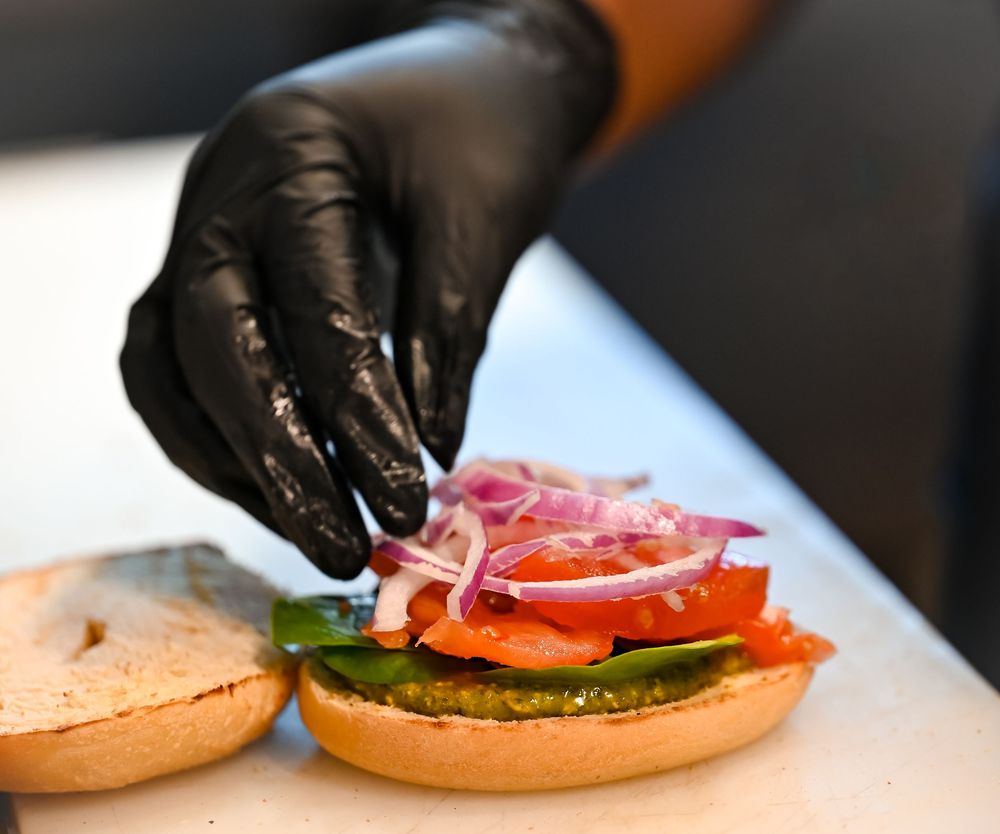 Chef in black gloves assembling a fresh bagel sandwich with smoked salmon, tomatoes, and red onions.