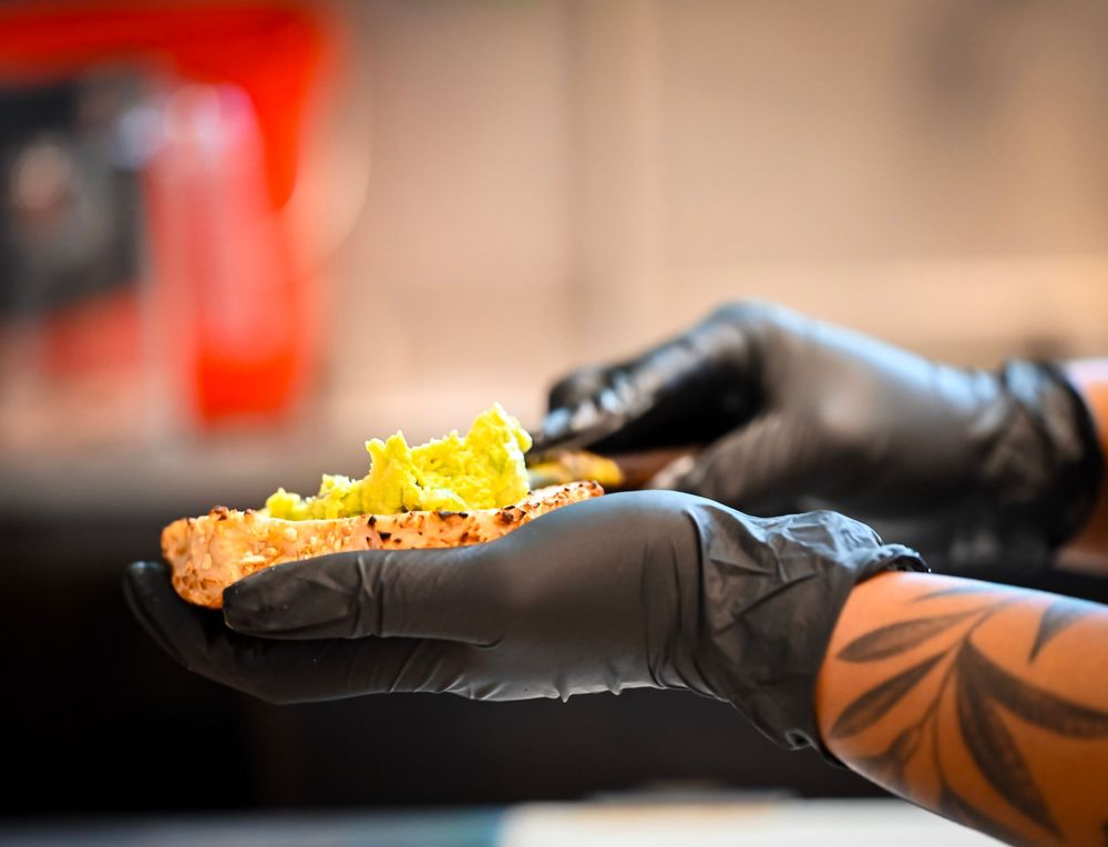 Tattooed hands in black gloves holding a seasoned burger patty topped with fresh lettuce.
