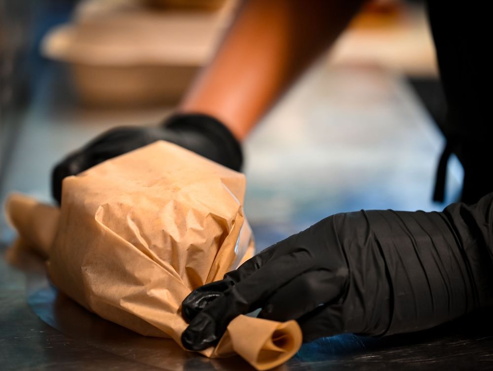 Chef in black gloves wrapping a burger in brown paper on a stainless steel kitchen counter.