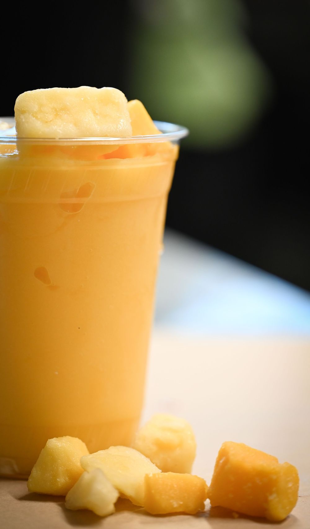 A refreshing mango smoothie in a clear cup topped with frozen mango chunks, served with diced fruit pieces on the side.
