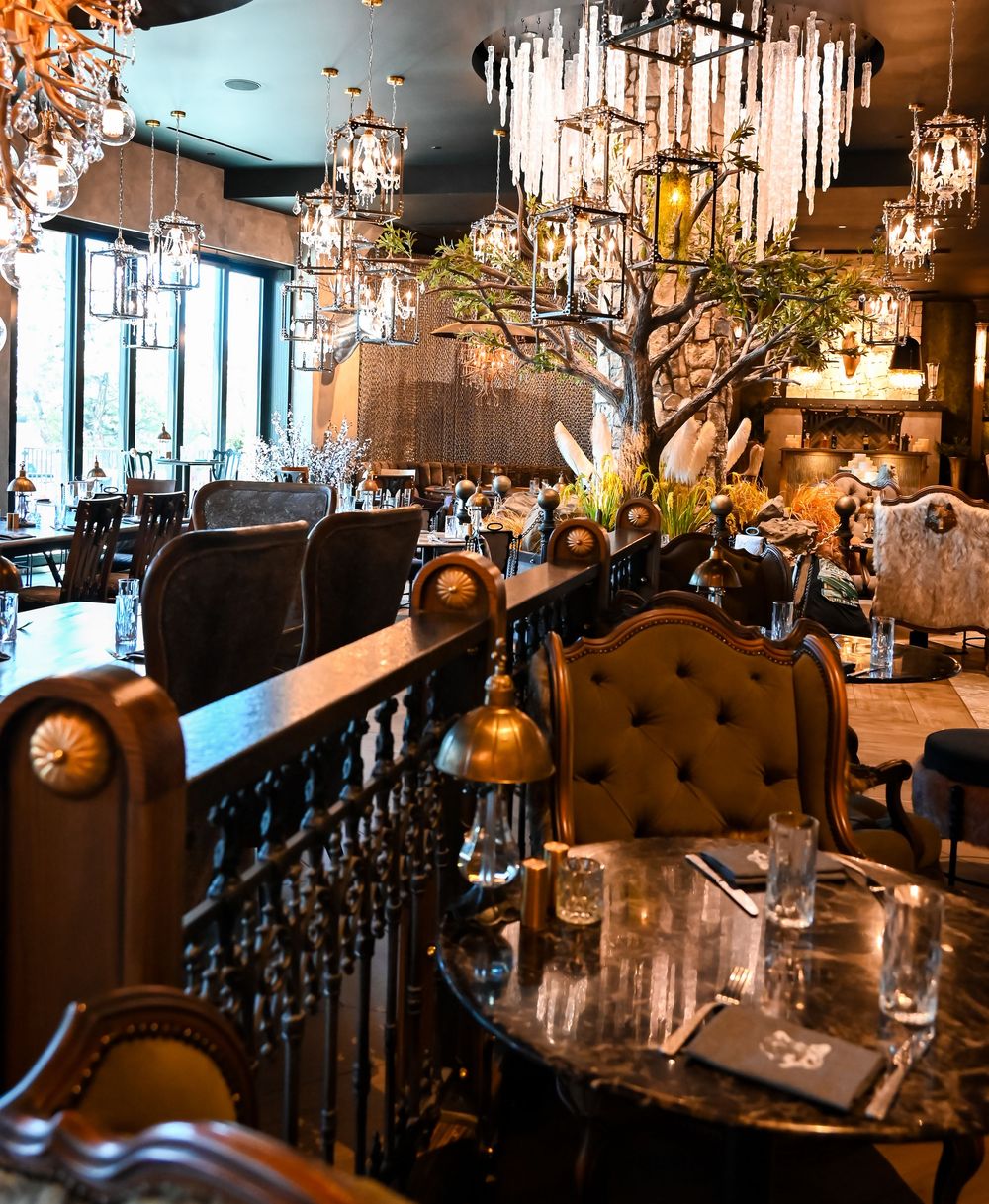Elegant restaurant interior with crystal chandeliers, tufted leather chairs, marble tables, and lush greenery decor.