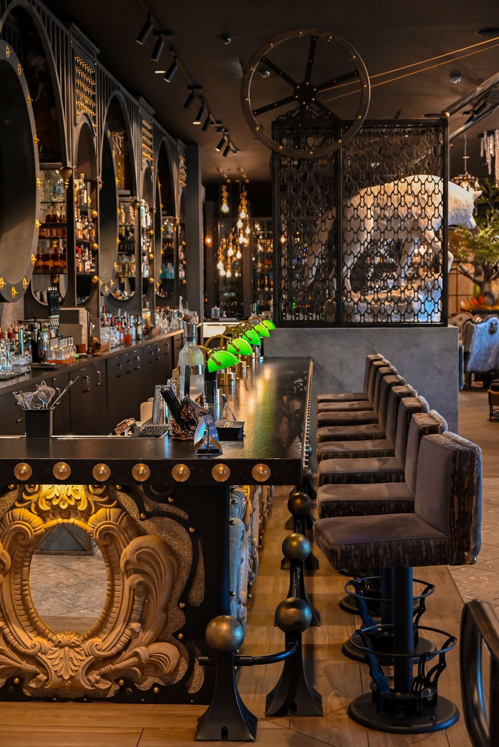Luxurious dark bar interior with ornate gold details, velvet bar stools, green bottle accents, and elegant metalwork decor.