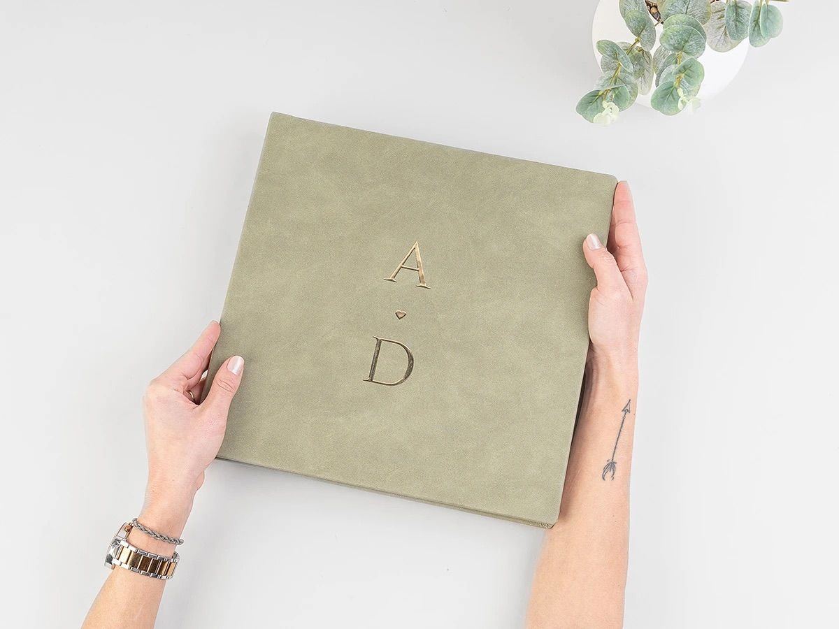 Hands holding a sage green suede wedding album embossed with gold initials A and D on a white background.