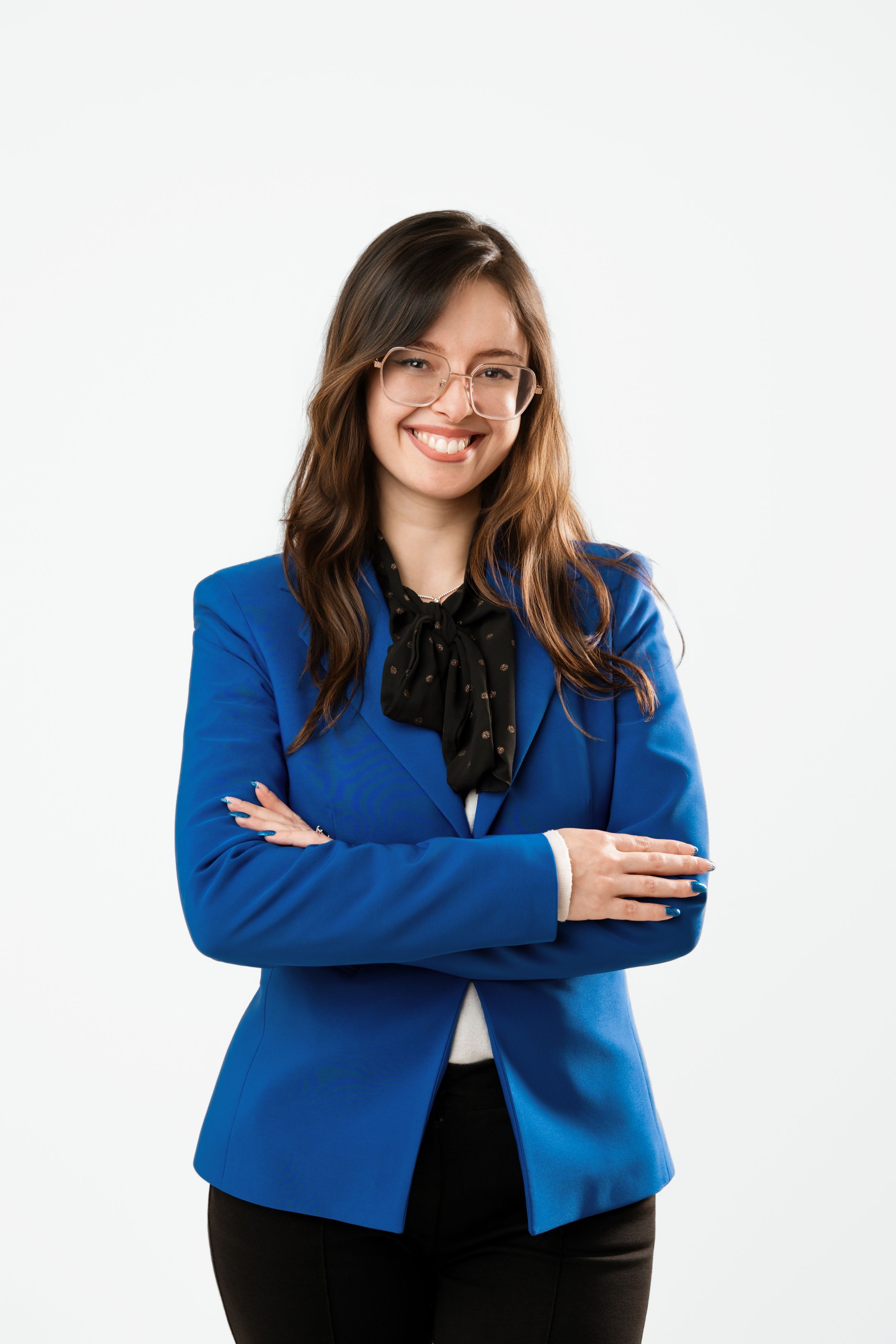 Smiling young woman in blue blazer and glasses, arms crossed, white studio background.