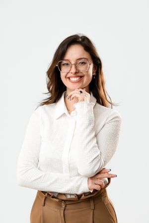 Cheerful woman in white blouse and glasses, hand on chin, wearing tan trousers, white background.