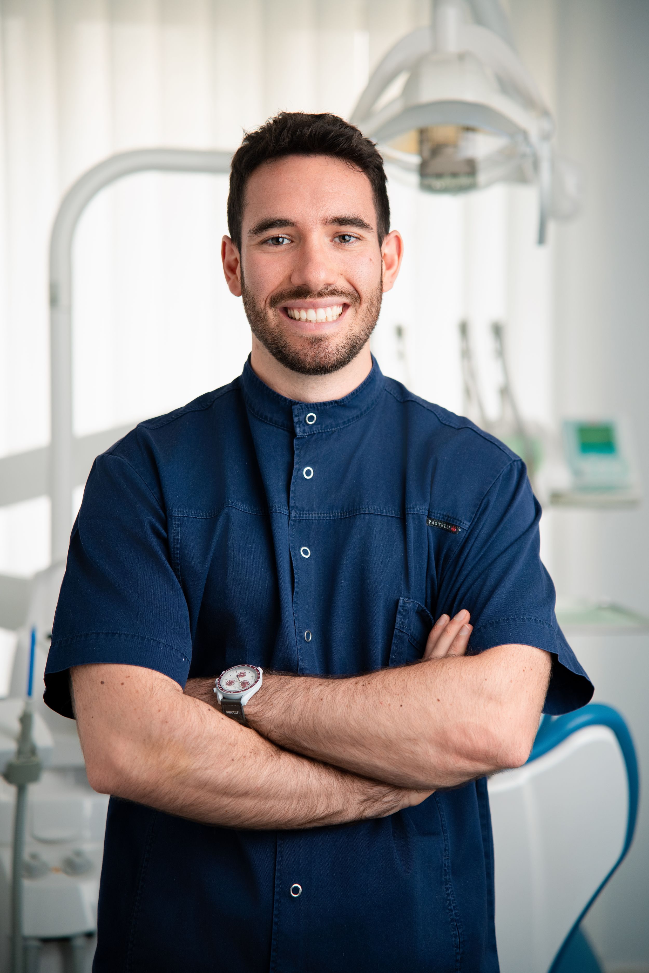 A smiling male dentist in navy scrubs stands with arms crossed in a modern dental clinic.