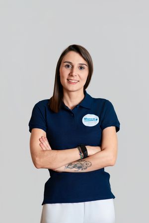 A smiling woman in a navy polo shirt with a name badge stands with arms crossed, showing a tattoo.