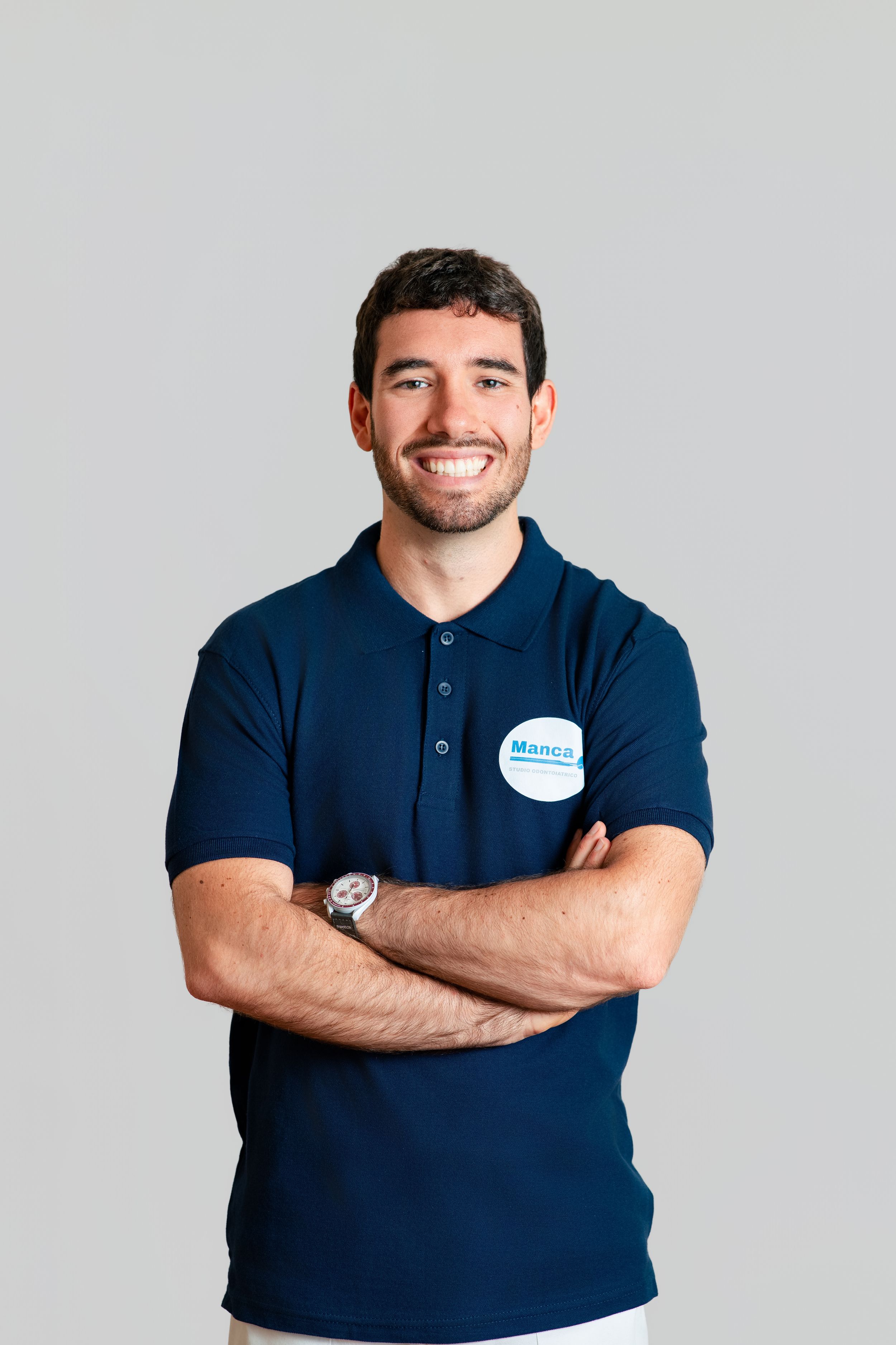 A smiling man in a navy polo shirt with a name badge stands confidently with arms crossed.
