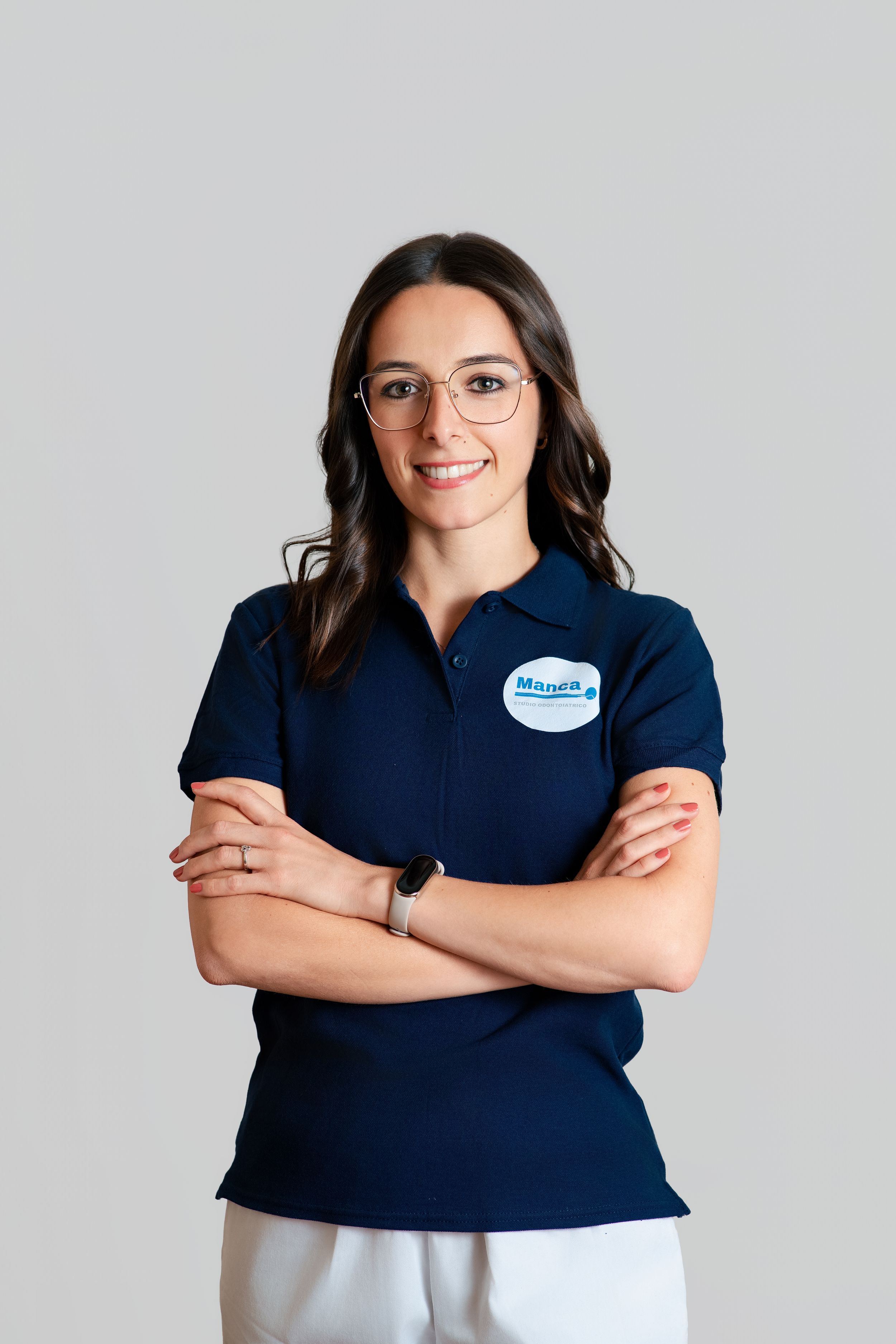 A smiling woman wearing glasses and a navy polo shirt with a name badge poses with arms crossed.