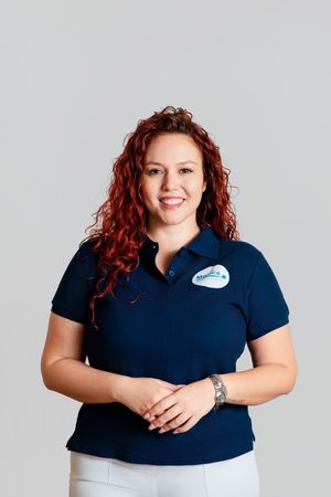 A cheerful woman with curly red hair in a navy polo shirt with a name badge smiles warmly.