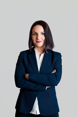 A professional woman in a navy blazer and white blouse stands with arms crossed against a gray background.