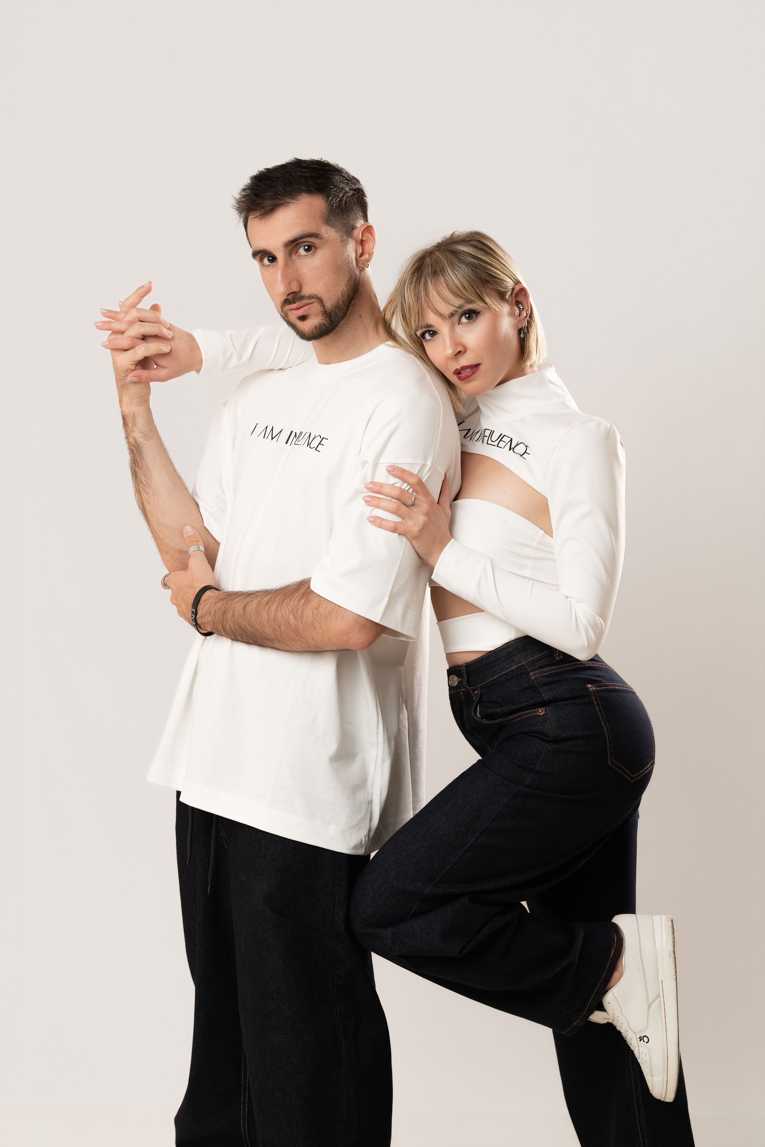 A man and woman in matching white graphic tees and black pants pose together against a light background.