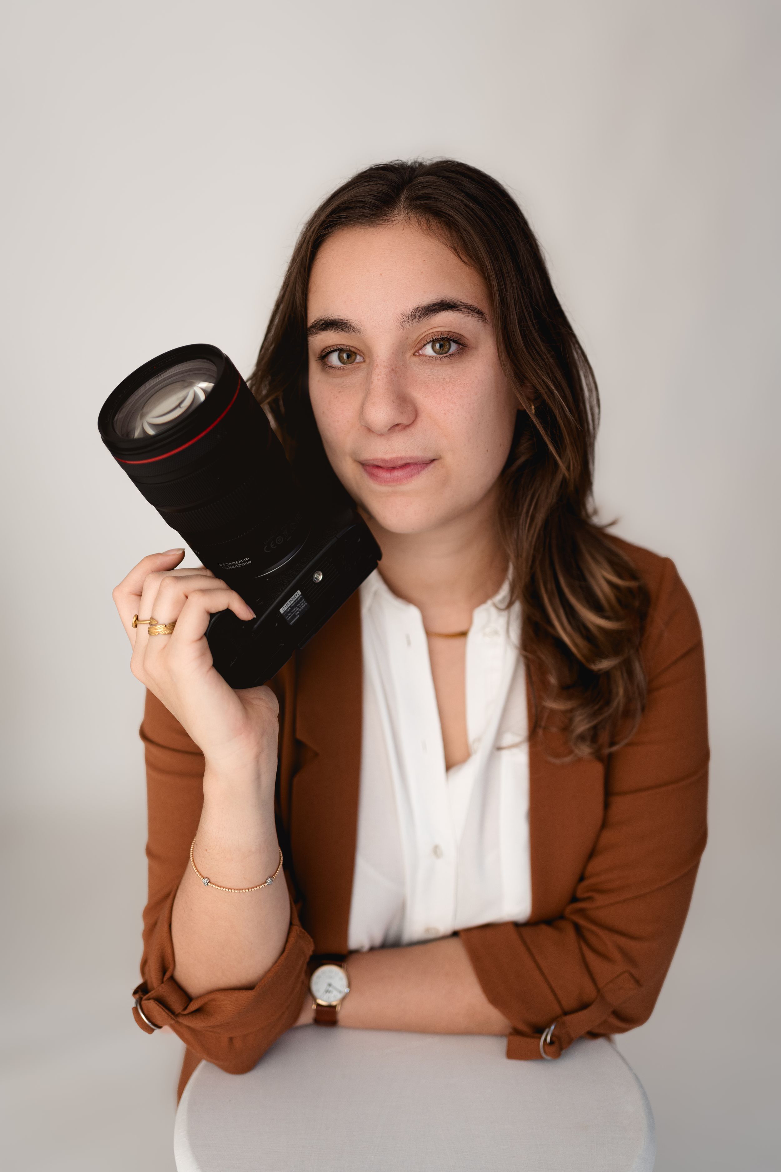 Professional female photographer poses holding a camera lens in a brown blazer in white studio.