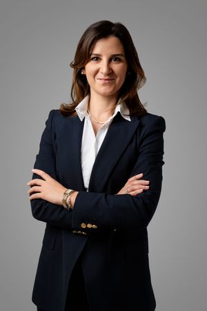 Professional woman in navy blazer and white shirt, arms crossed, smiling on gray background.