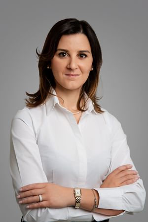 Professional woman in white shirt with arms crossed, wearing gold watch, gray background.