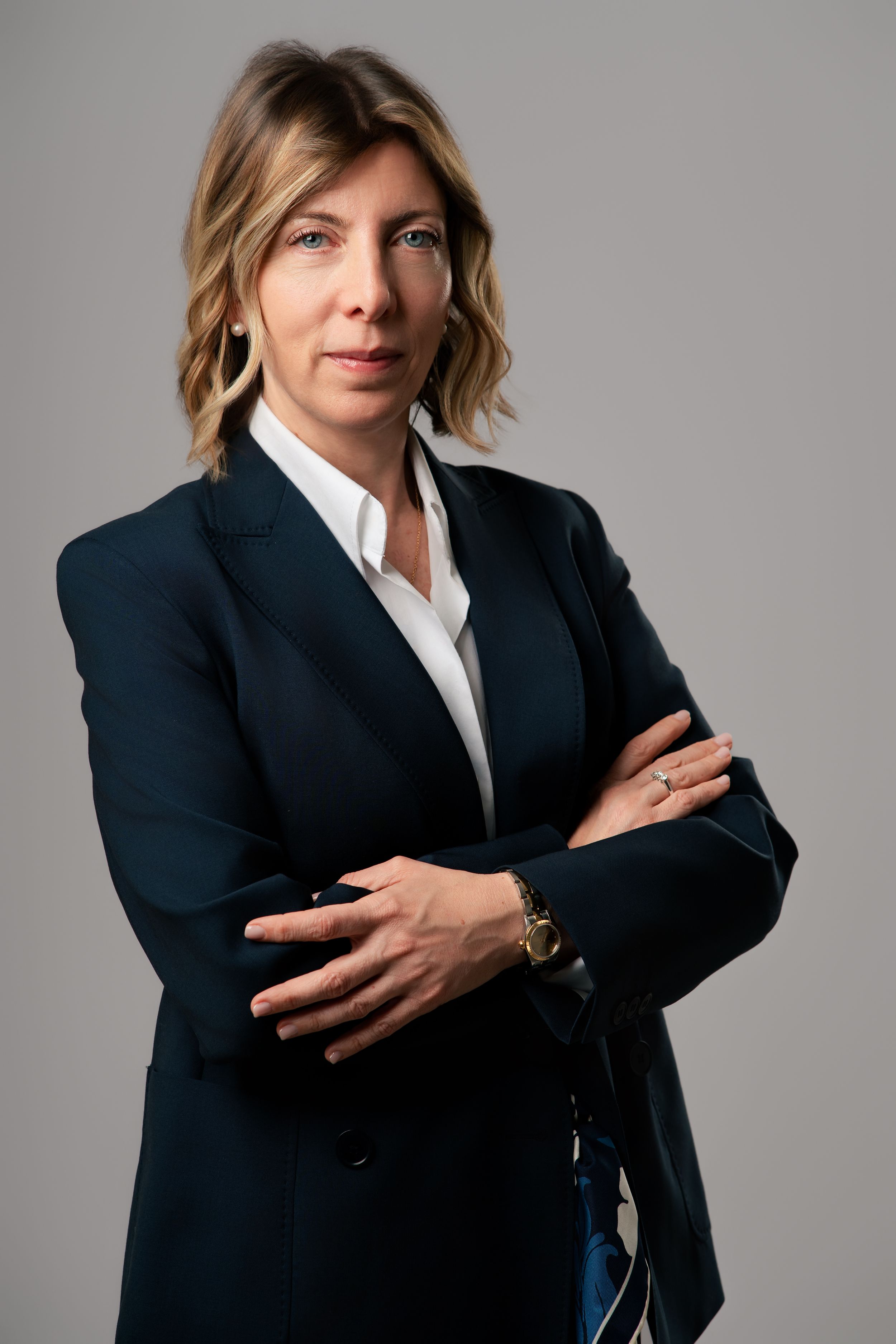 A serious businesswoman in a navy blazer and white shirt stands with arms crossed against a gray background.