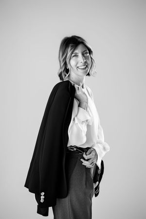 Smiling woman in white blouse holding black blazer over shoulder, black and white professional portrait photo.