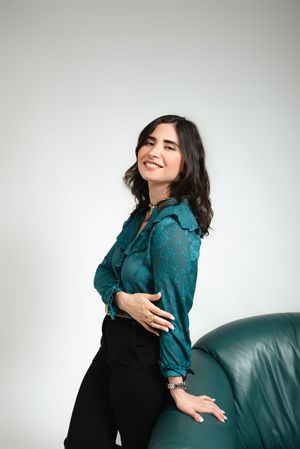 Woman in teal patterned blouse posing beside green leather chair, smiling over shoulder.