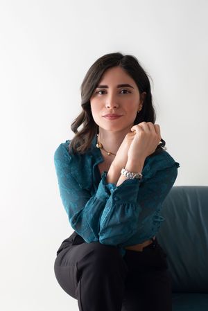 Dark-haired woman in teal blouse seated on sofa, hands clasped, looking up thoughtfully.