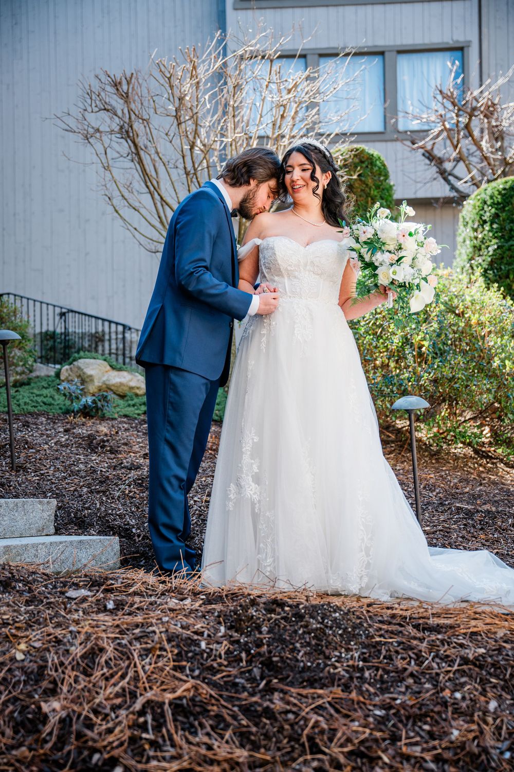 A groom in a blue suit kisses his bride holding a white bouquet outdoors in a garden setting.