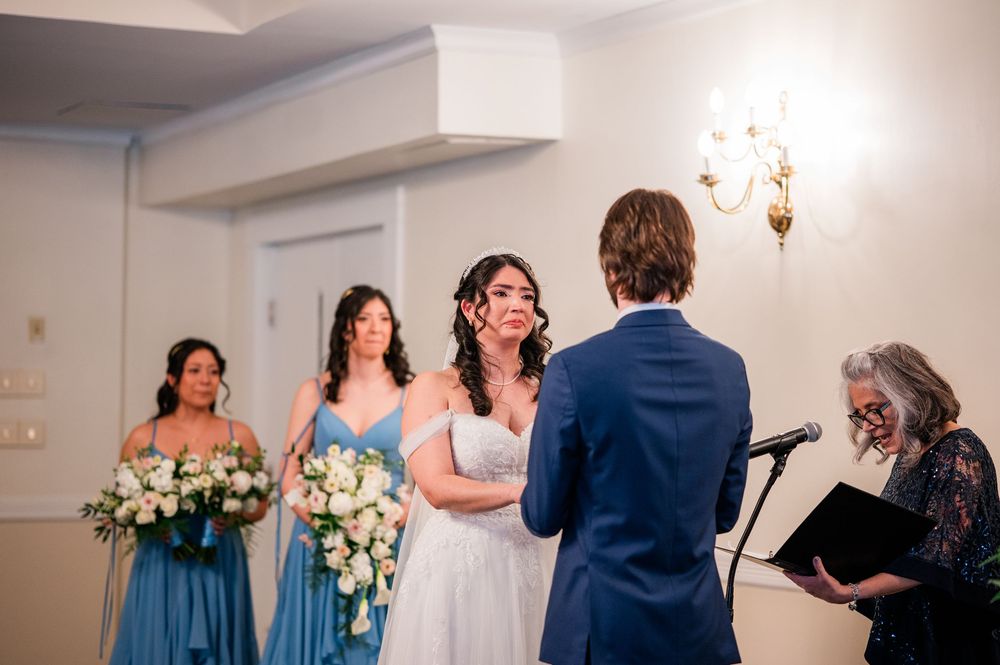 Bride smiling at groom during indoor wedding ceremony with bridesmaids in blue dresses holding white floral bouquets.