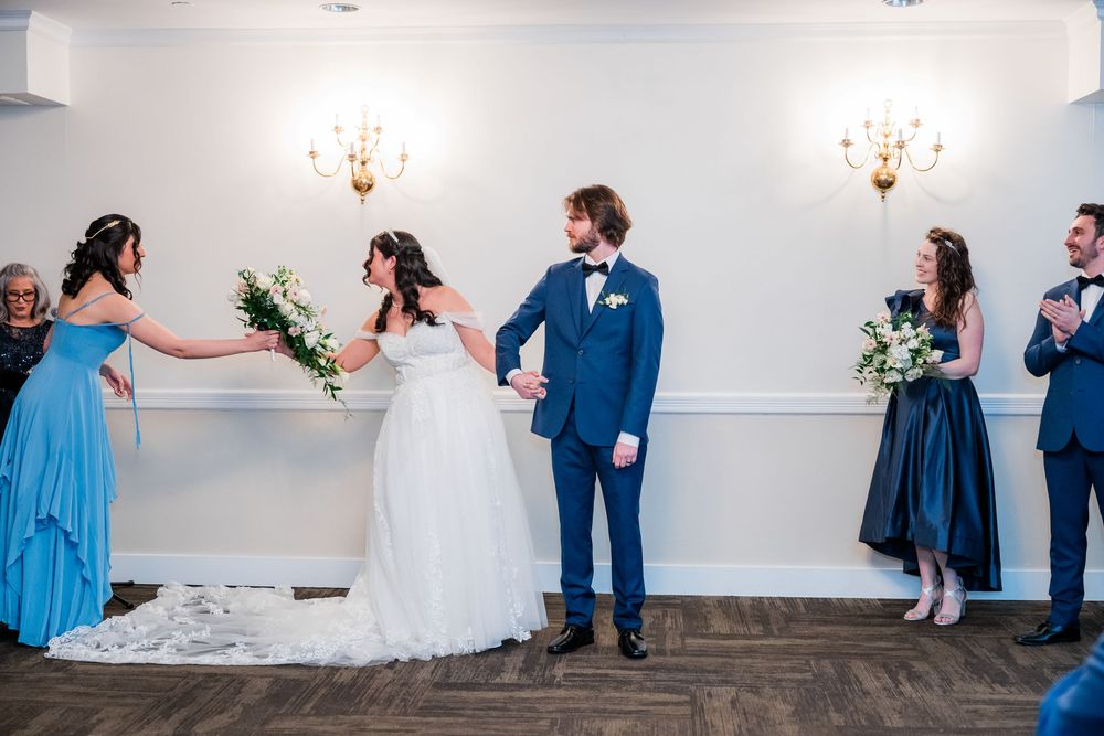 Bride and groom hold hands during indoor wedding ceremony with bridal party in blue and navy formal attire.