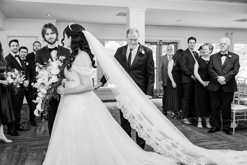 Black and white wedding ceremony photo of bride in veil gown meeting groom at altar with guests standing nearby.