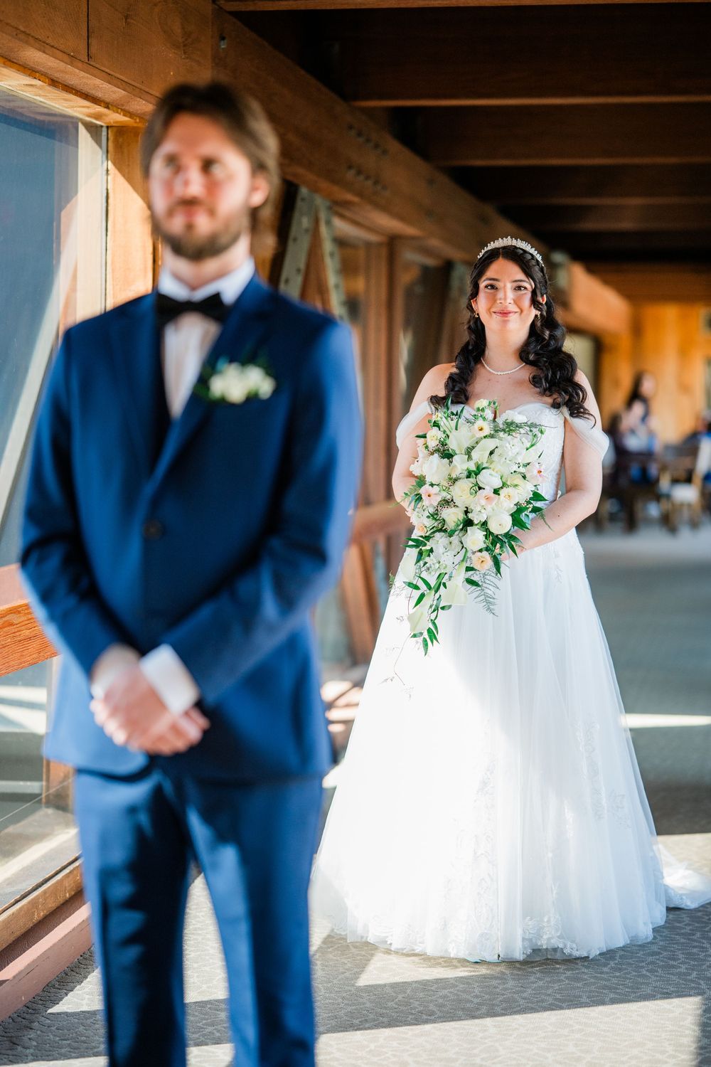 A bride in a white lace ball gown holds a bouquet while her groom stands in the foreground during first look.