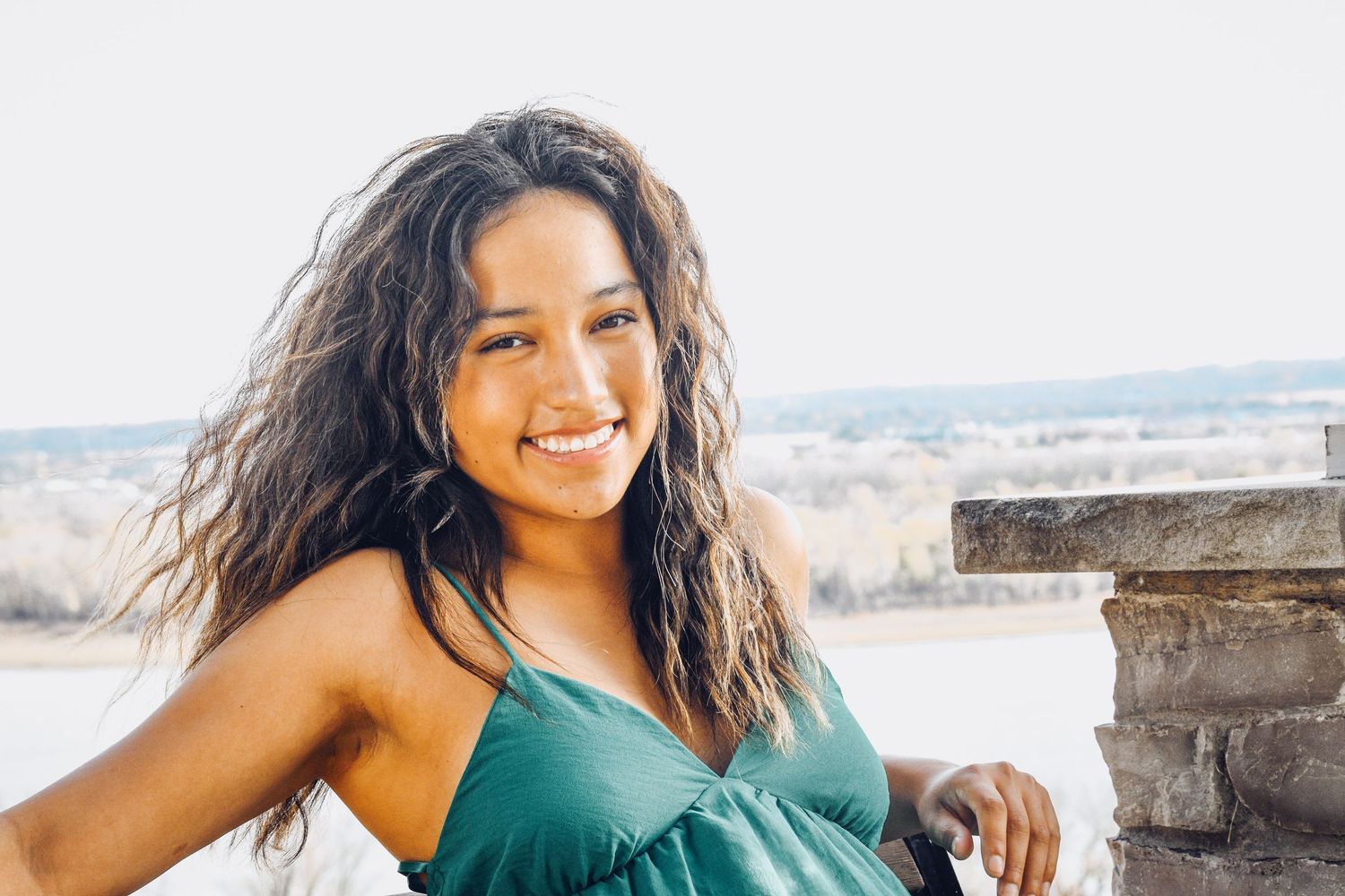 Young woman with curly hair smiling outdoors in a green top, with a scenic landscape in the background.