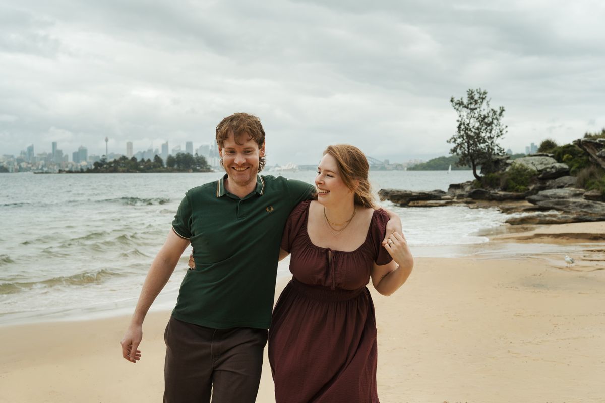 Couple walking and laughing on a sandy beach with city skyline and rocky shore in the background in Milk Beach engagement photoshoot