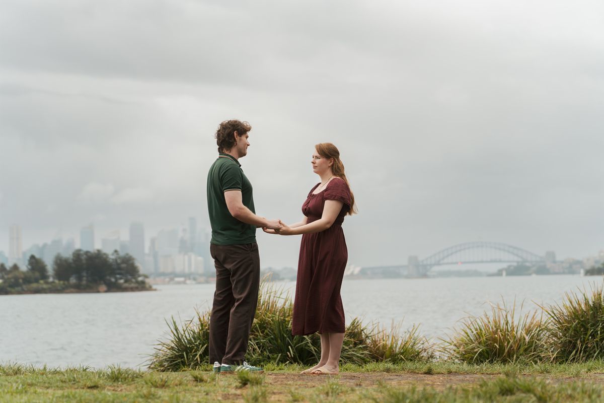Couple holding hands by Sydney Harbour with bridge backdrop on overcast day during an engagement photoshoot in Milk beach