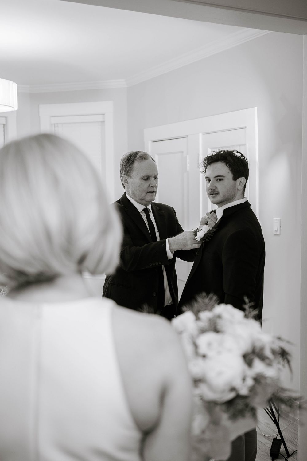 Older man helping groom adjust boutonniere while bride holds bouquet in black and white wedding photo.