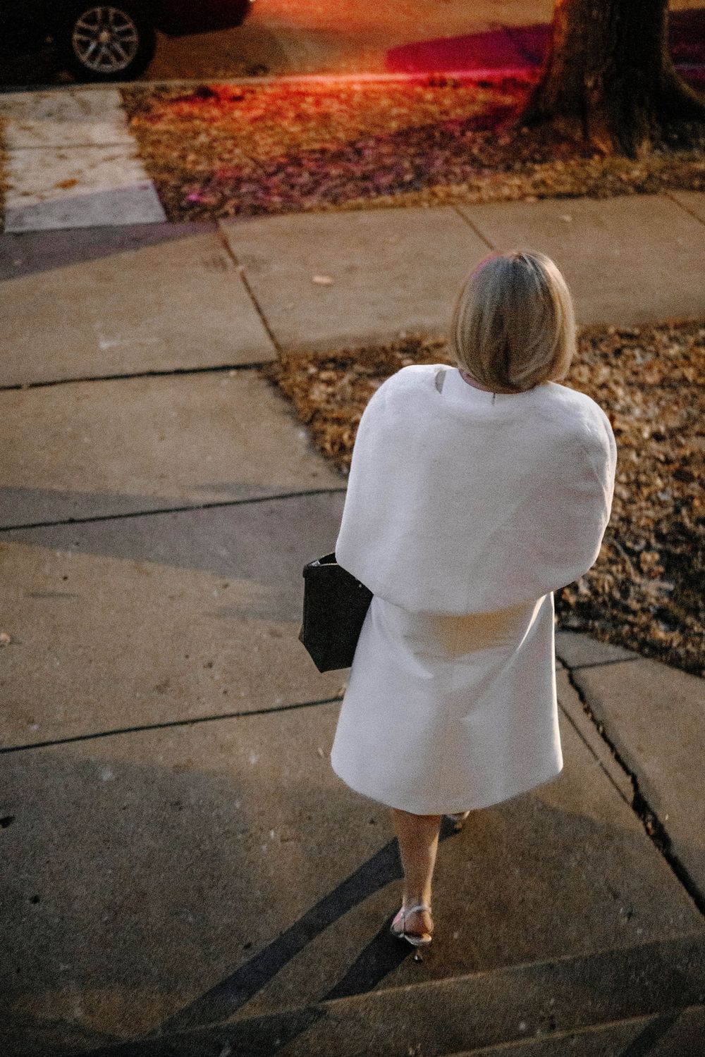 Elegant woman in white cape coat holding black clutch walks along a sidewalk at dusk.