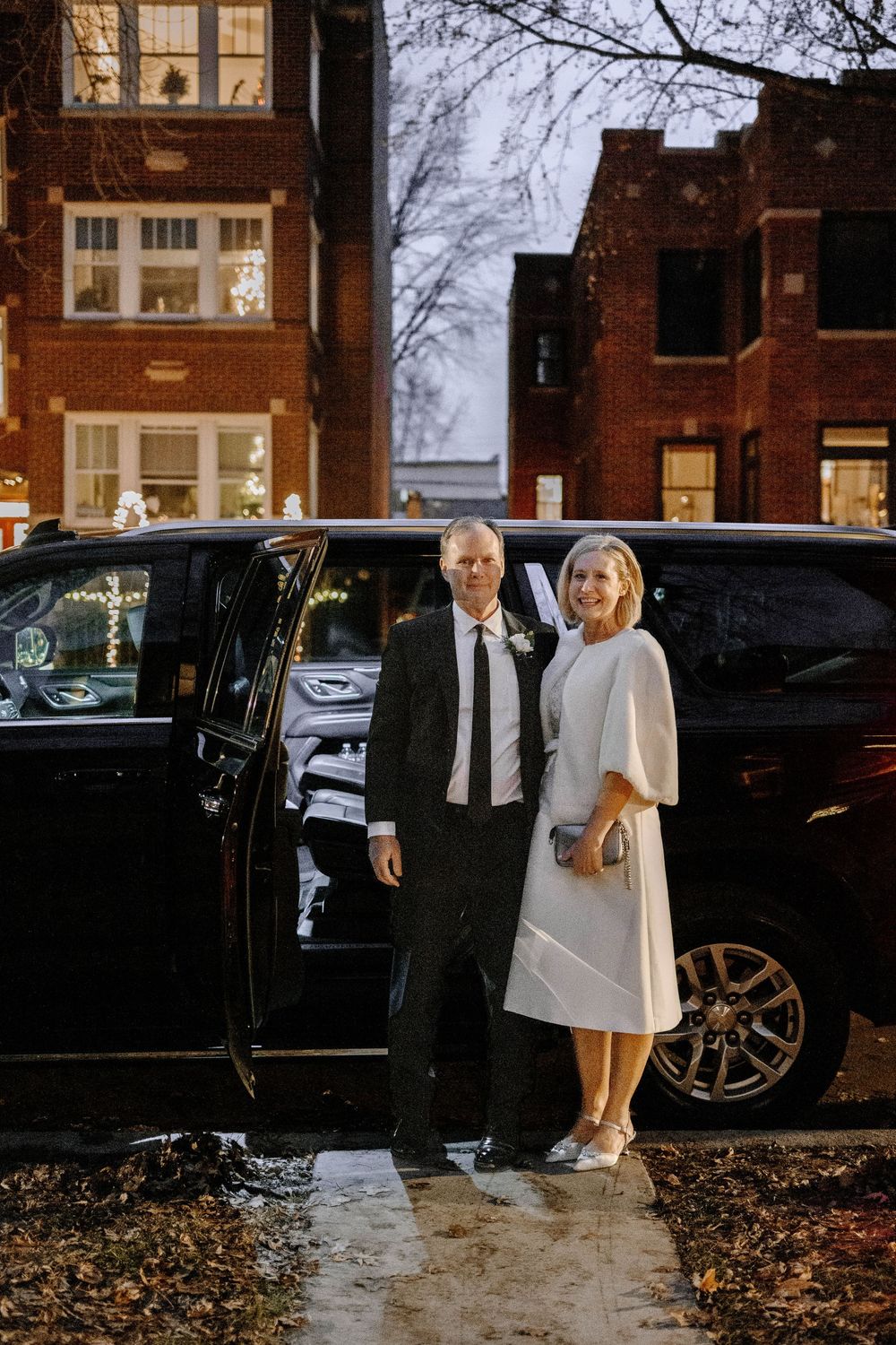 Elegant couple in wedding attire posing beside a black luxury SUV on a city street at night.
