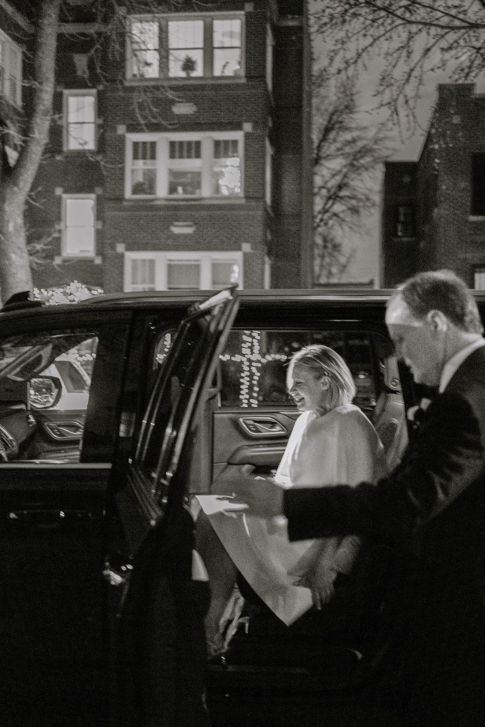Black and white photo of a bride in white gown being helped into a luxury car by a man in a suit.