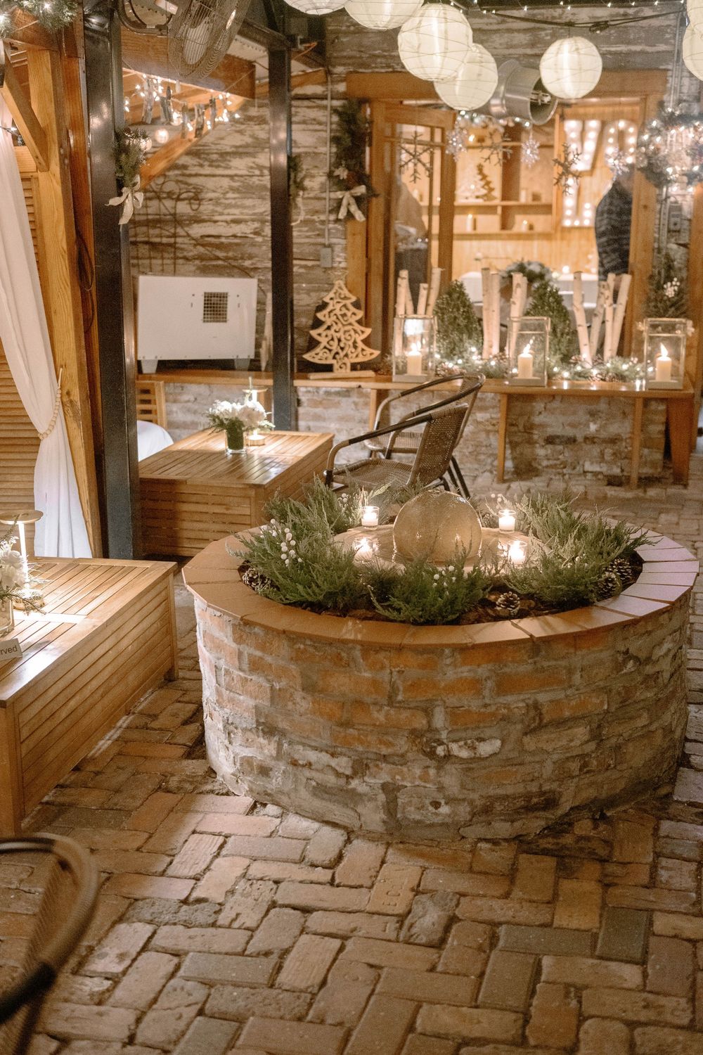 Rustic wedding venue with stone fire pit centerpiece, candles, greenery, string lights, and paper lanterns creating warm ambiance.