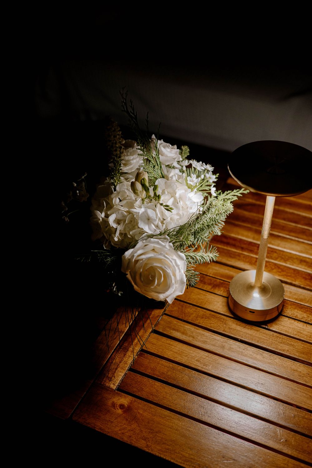 White roses and hydrangea bouquet beside a gold lamp on a wooden slatted table in dramatic lighting.