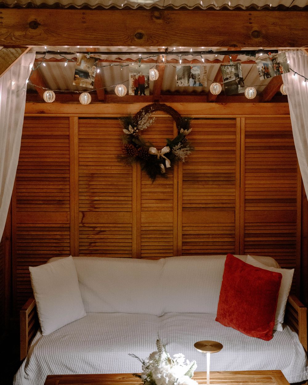 Cozy wedding lounge nook with white cushioned sofa, red pillow, floral wreath, fairy lights, and shiplap wood wall backdrop.