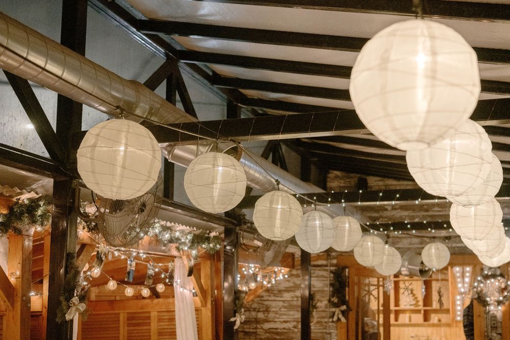 White paper lanterns of varying sizes hanging from rustic wooden beam ceiling with warm string lights at wedding venue.