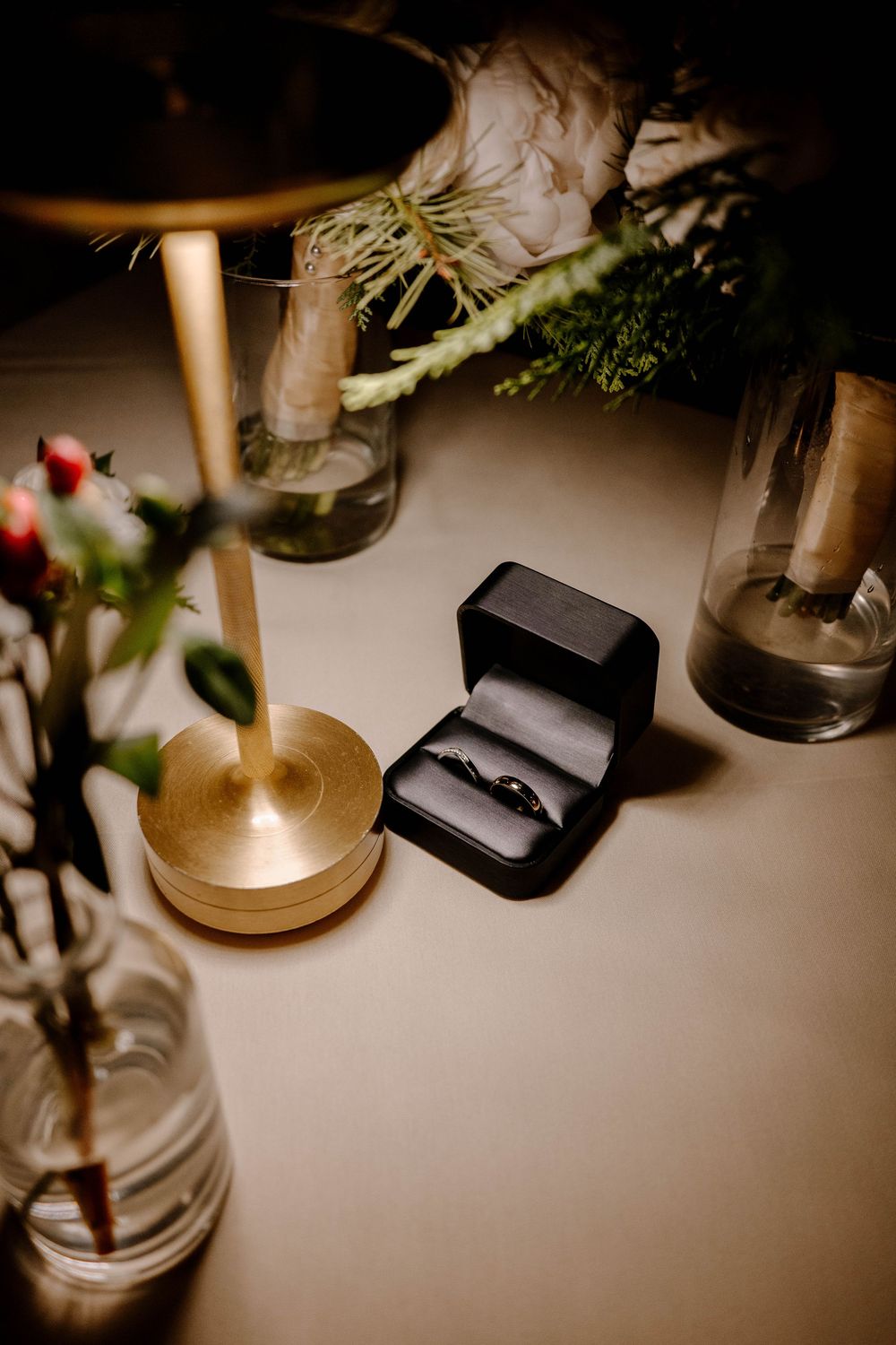 Black velvet ring box with wedding bands displayed on a table with floral arrangements and gold candlestick.