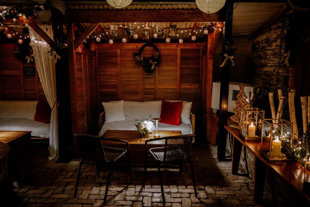 Cozy outdoor lounge area with string lights, wreath, wood paneling, white sofa, red pillows, and candles at night.