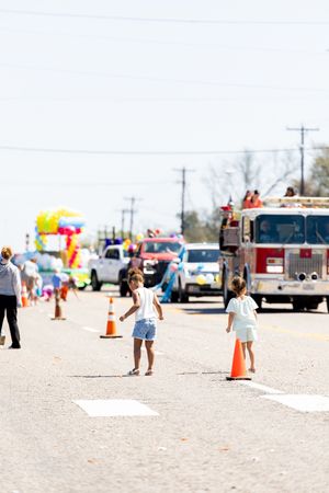 Two children stand on a street during a sunny parade with a fire truck and colorful floats in the background.