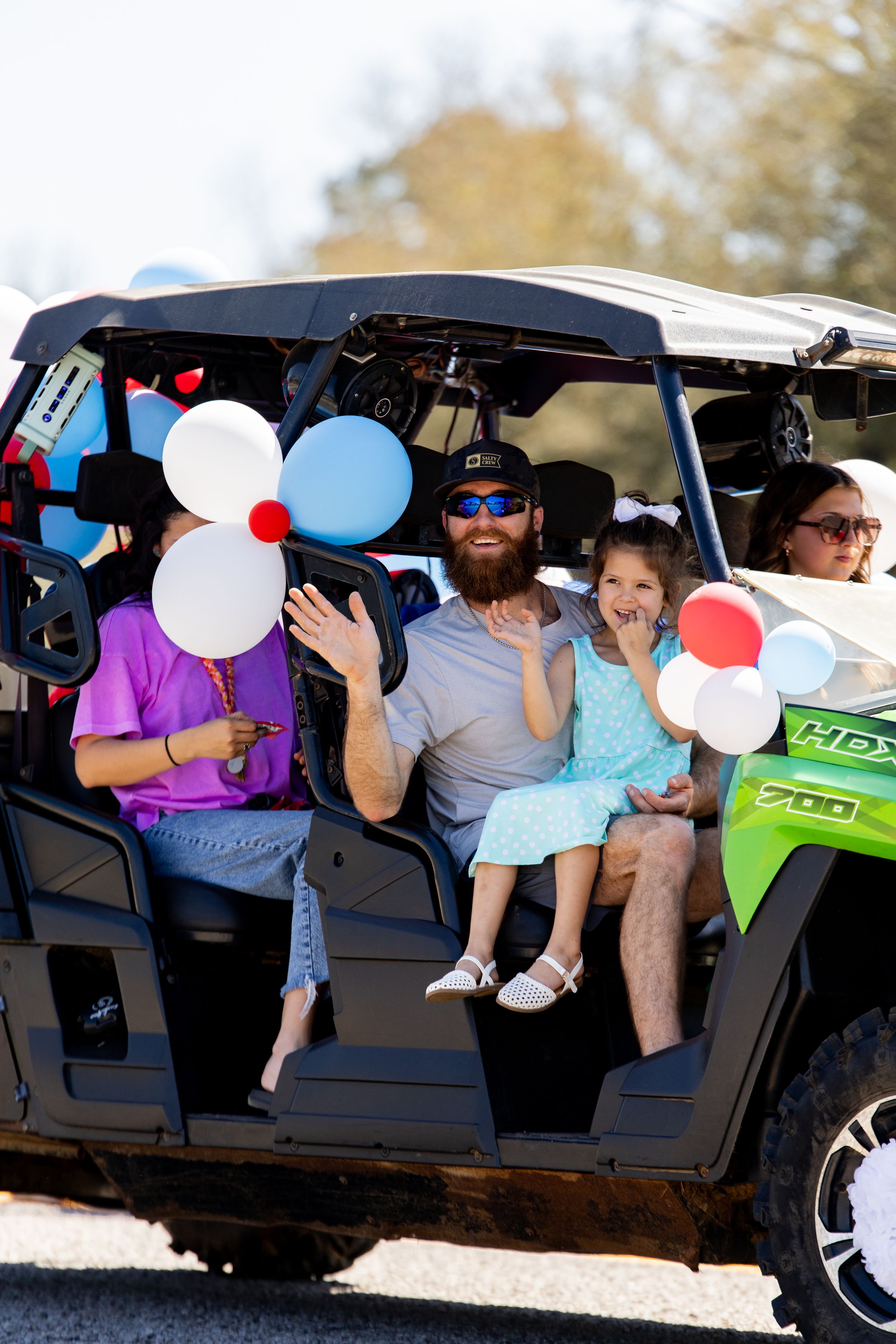Family riding in a green UTV at a parade, holding red, white, and blue balloons while waving.