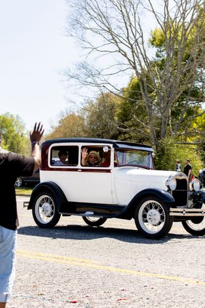 Vintage white antique car driving in a parade with passengers waving to crowd on a sunny day.