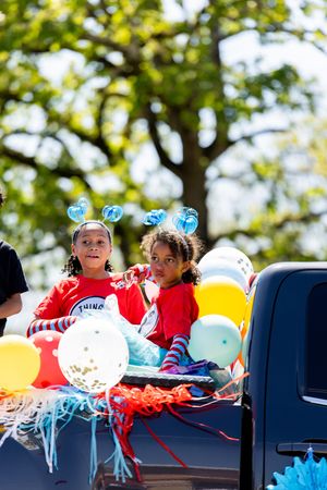 Two girls in red Thing costumes with blue heart headbands ride a colorful balloon-decorated parade float.
