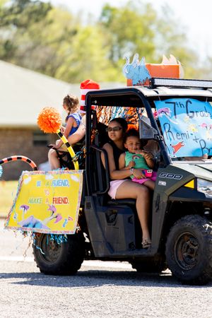 Women and child ride in decorated UTV during school parade with cheerleader and colorful signs visible.