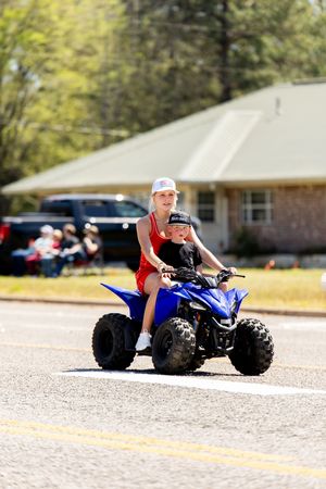 Two children ride a blue ATV quad bike down a sunny parade street, with a brick house visible in the background.