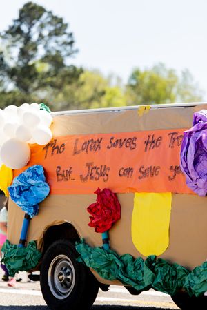 Colorful parade float decorated with paper flowers and balloons displaying a religious banner referencing The Lorax and Jesus.