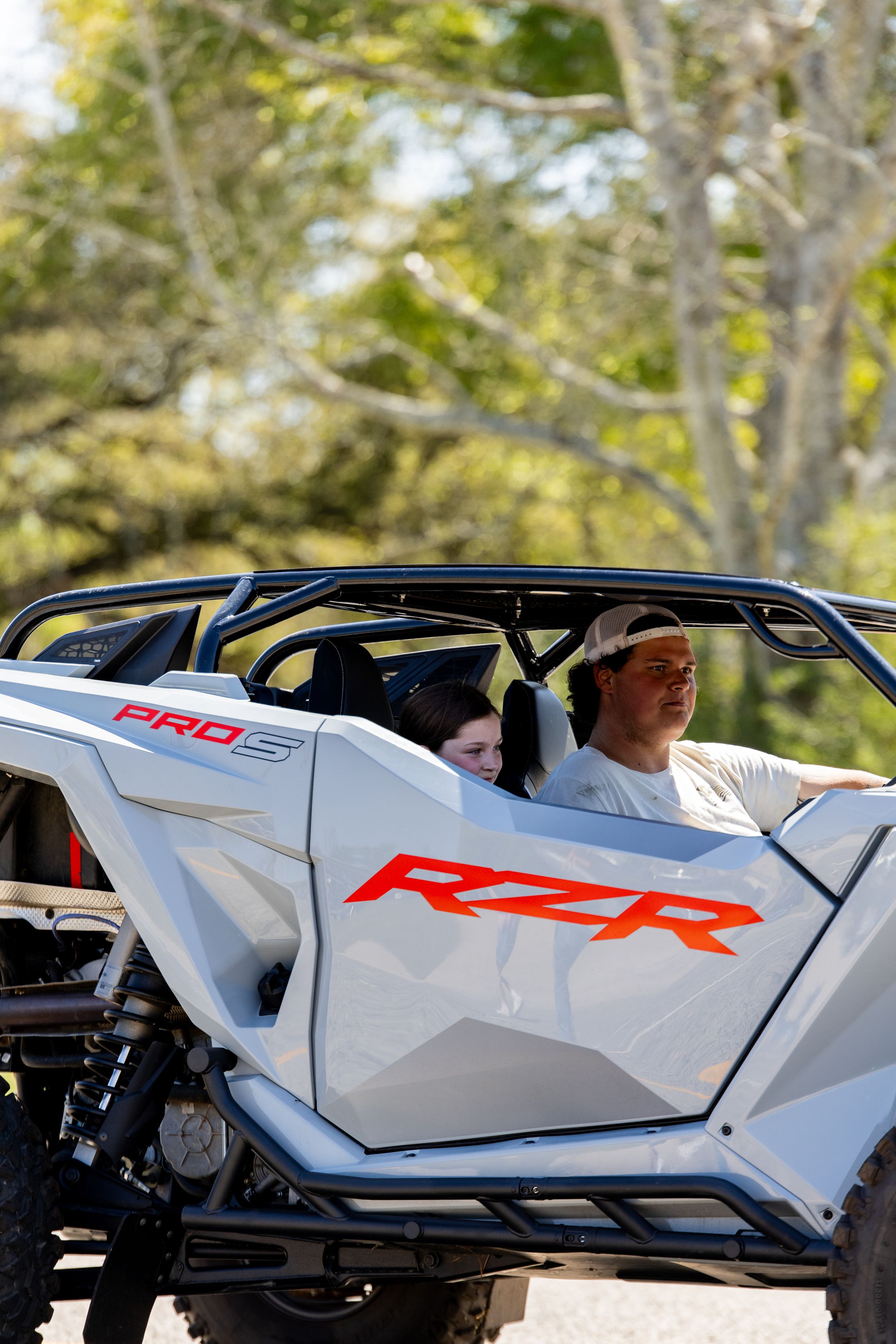 Two people riding in a white Polaris RZR Pro R off-road UTV through a wooded trail outdoors.