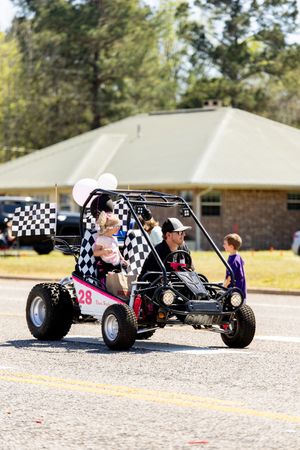 Man drives a custom go-kart with checkered flags in a small town parade, with children riding along.